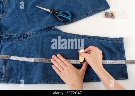 Le mani caucasiche della donna misurano la lunghezza desiderata con un nastro di misurazione su un tavolo bianco. Accorciare i jeans con forbici e spilla per cucire. FAI DA TE. Foto Stock