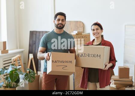 Ritratto di giovane coppia che tiene scatole e sorridendo alla macchina fotografica mentre si muove in nuova casa Foto Stock