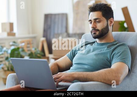 Giovane uomo con la bearded che riposa su poltrona con computer portatile nella stanza a casa Foto Stock