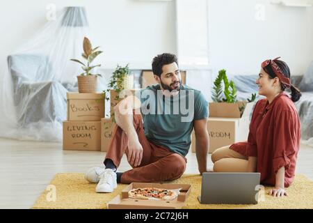 Giovane coppia seduta sul pavimento con un computer portatile che parla l'un l'altro e che mangia la pizza nella loro nuova casa Foto Stock
