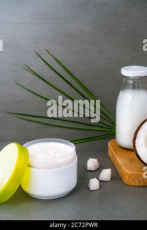 Crema spa aperta per la pelle del corpo e i capelli con olio di cocco, noci di cocco fresche, foglia di palma e bottiglia con burro di cocco su sfondo grigio scuro. Naturale Foto Stock