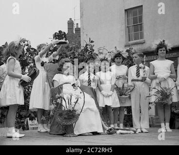 Anni '50, storica, in un centro cittadino, fuori su una piattaforma di legno, una giovane ragazza seduta in un sedile che viene incoronata la 'May Queen', con altri bambini piccoli in piedi accanto a lei, Inghilterra, Regno Unito. Foto Stock