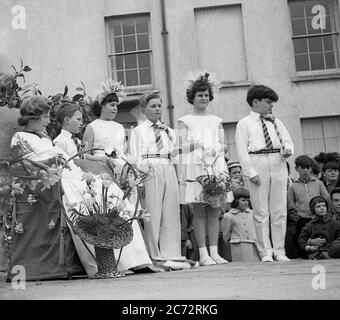 Anni '50, storica, fuori in un centro di mercato, su una piattaforma di legno, una giovane ragazza seduta in un posto pronto ad essere incoronata come la 'May Queen', con altri bambini piccoli in piedi accanto a lei, Inghilterra, Regno Unito. Foto Stock