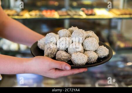 Vista selettiva messa a fuoco delle mani della donna che tengono un vassoio di deliziosi tartufi di cioccolato bianco, pasticceria mostra frigorifero sfocato sullo sfondo. Foto Stock