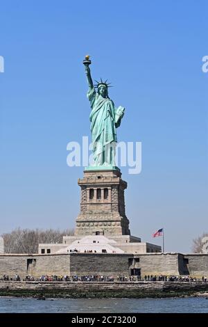 La Statua della libertà, Liberty Island, Manhattan, New York. I visitatori possono essere visti passeggiando intorno alla base della statua. Cielo azzurro chiaro, bandiera americana. Foto Stock