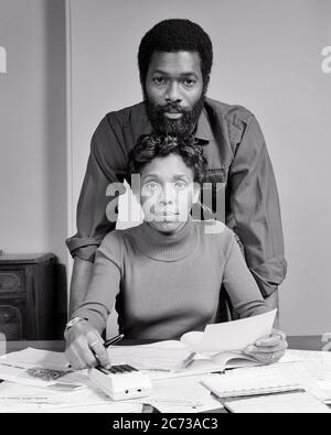 1970 COPPIA AFROAMERICANA GUARDANDO LA MACCHINA FOTOGRAFICA UOMO IN PIEDI DIETRO LA DONNA SEDUTA AL TAVOLO CON CARTE BOLLETTE MANO SU CALCOLATRICE - S21331 HAR001 HAR001 CALCOLATRICE HARS VISO BILANCIO STILE COMUNICAZIONE TEAMWORK STILE DI VITA DONNE SPOSATE SPOSATE SPOSATE MARITI HOME VITA COPIA SPAZIO AMICIZIA MEZZA LUNGHEZZA DONNE UOMINI RISCHIO ESPRESSIONI B&W PARTNER OCCHIO CONTATTO OBIETTIVI AFROAMERICANI AFROAMERICANI ETNIA NERA INNOVAZIONE CAPELLI VISO CONNESSIONE BARBE COOPERAZIONE MID-ADULT UOMO MID-ADULT DONNA SOLUZIONI TOGETHERNESS MOGLI NERO E BIANCO HAR001 TLENECK VECCHIO STILE SORRIDENTE Foto Stock