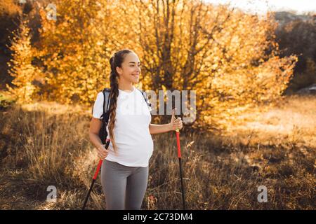 Donna incinta nordica Walking nella foresta autunnale con zaino e bastoni da trekking. Stile di vita sano e attivo nel tempo di maternità. Esercizi di gravidanza per il benessere. Foto Stock