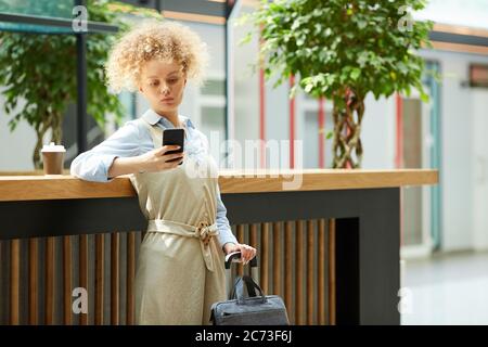 Giovane donna d'affari in piedi con valigia e utilizzando il telefono cellulare mentre si beve caffè in caffetteria Foto Stock