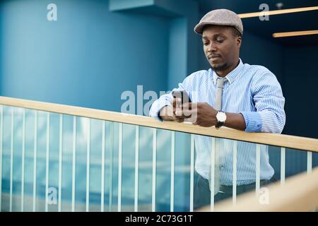 Giovane africano in berretto in piedi nel corridoio dell'ufficio e digitando un messaggio sul telefono cellulare Foto Stock