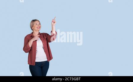 Donna caucasica senior con capelli biondi che punta allo spazio libero blu studio vicino a lei con dito indice Foto Stock