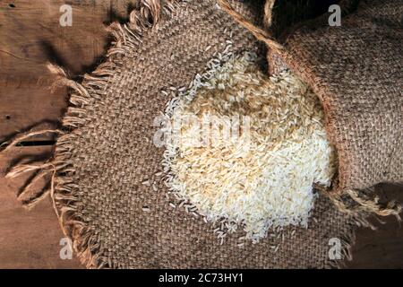 Riso fuori da piccolo sacco isolato su sfondo di legno. Riso fresco in sacco su fondo testurizzato di legno. Chiudi su grano di riso crudo su panno di sacco. Foto Stock