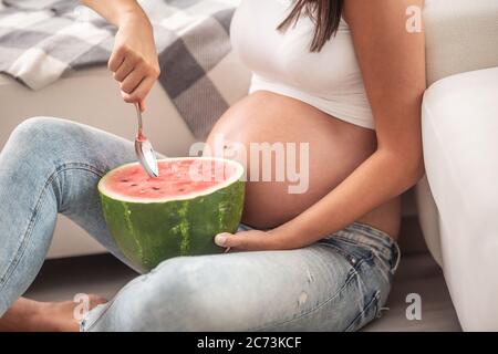 Una donna incinta seduta sul pavimento mangia un anguria con un cucchiaio. Foto Stock
