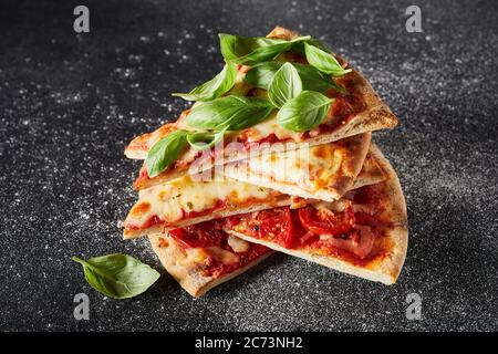 Fette di pizza Margherita appena sfornate impilate una sull'altra e ricoperte di foglie di basilico fresco su sfondo scuro di ardesia con farina Foto Stock