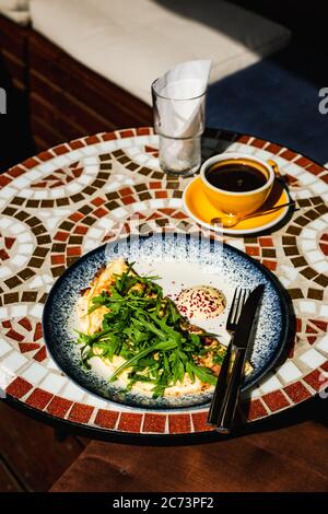 Una colazione composta da una frittata con rucola o insalata di razzo, una tazza gialla di caffè nero, su un tavolo in pietra a mosaico di un caffè in una giornata di sole Foto Stock