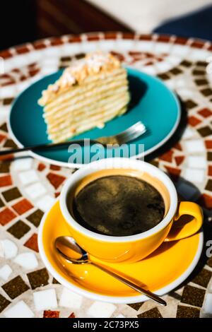 Una fetta di torta napoleone su un piatto blu e una tazza gialla di caffè nero in una giornata di sole su una terrazza di un caffè, tavola di pietra mosaico Foto Stock