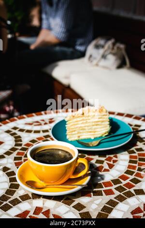 Una fetta di torta napoleone su un piatto blu e una tazza gialla di caffè nero in una giornata di sole su una terrazza di un caffè, tavola di pietra mosaico Foto Stock