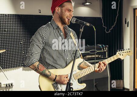 il giovane chitarrista caucasico suona contemporaneamente la chitarra e canta una canzone in studio di registrazione. bel ragazzo in red hat cantando emotivamente canzone, appassionato di mu Foto Stock
