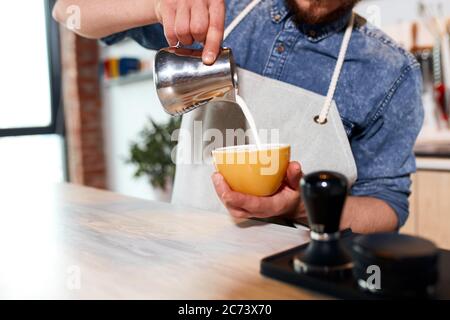 La quantità di latte caldo che viene versato nella tazza di caffè, il barista professionista tiene in mano una tazza di caffè bellissima e aggiunge latte cotto al vapore nel latte Foto Stock