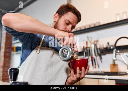 Il barman di talento e sicuro mantiene una magnifica tazza rossa con latte caldo e gustoso, versa il latte in una tazza di caffè, in piedi dietro il bancone, in una camera ben arredata Foto Stock