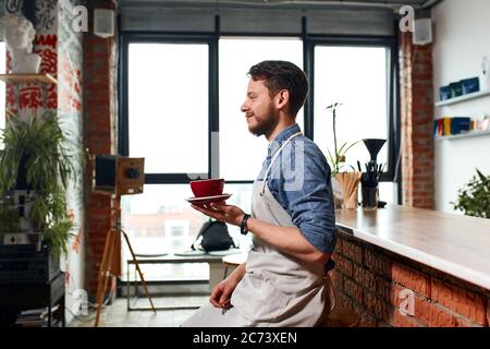 Ottimista barista maschile allegro, si siede vicino al bancone bar, tiene bella tazza con caldo gustoso americano, guarda da parte con viso positivo, sorridendo gentilmente, Foto Stock