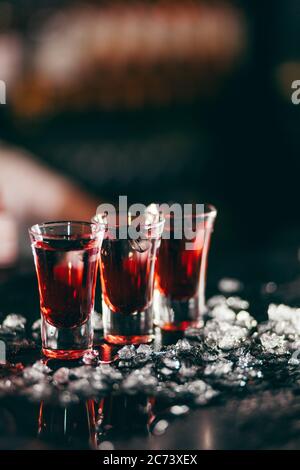 Tre colpi rossi di alcool in linea circondati da pezzi di ghiaccio, isolati su sfondo scuro sfocato. Concetto di alcol, alcolici per una buona notte Foto Stock