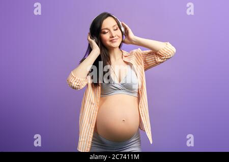 Donna in gravidanza felice sentirsi sano ascoltare musica in cuffia, ballare in studio su sfondo viola Foto Stock