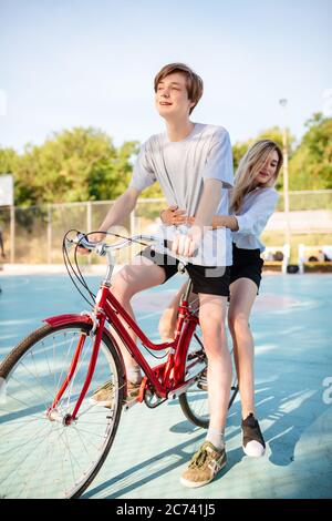 Sorridente ragazzo e bella ragazza con capelli biondi in bicicletta felicemente guardando da parte mentre si passa il tempo sul campo da basket. Giovane coppia carina che cavalcano rosso Foto Stock