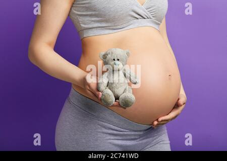 Donna incinta che tiene l'orso peloso giocattolo, vicino al suo grande ventre, isolato su viola, primo piano. L'orso giocattolo come simbolo della maternità di soom e della nascita del bambino Foto Stock