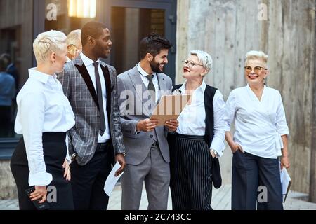 Gruppo di uomini d'affari si spostano all'aperto. Nel centro brunetto caucasico uomo bearded in elegante vestito grigio sorriso ampiamente, tenere papier. Donna vicino sorridere labbra rosse, Foto Stock