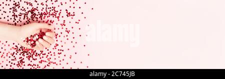 Bellissima femmina mano azienda rosso cuore di lamina con vibrante rosso spumante a forma di cuore coriandoli versata su sfondo rosa. Lay piatto. L amore e la celebratio Foto Stock