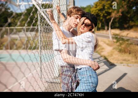 Ragazzo pensivo in piedi e abbracciando bella ragazza con capelli biondi. Bella ragazza in jeans e nero tappo in piedi con ragazzo sul banco di basket Foto Stock