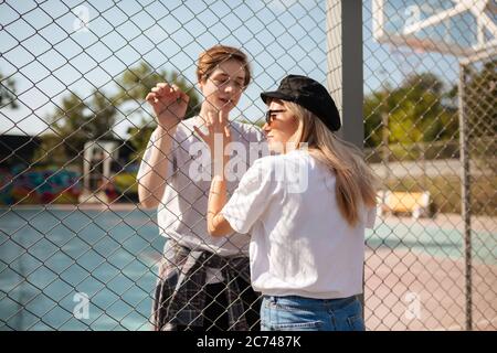 Giovane bella coppia in piedi e guardando l'un l'altro attraverso recinzione mesh. Ragazzo pensieroso in piedi sul campo da pallacanestro e guardando bella ragazza Foto Stock