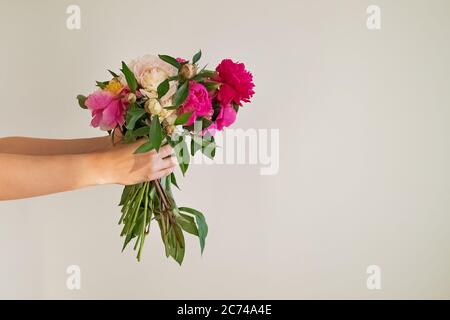Mani femminili che tengono un bouquet di belle peonie contro la parete bianca Foto Stock