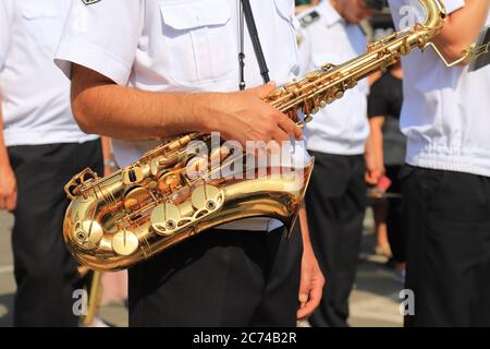 Un musicista maschile in una camicia bianca tiene un grande e bel sassofono dorato in una colonna di musicisti militari, primo piano. Strumento Jazz Foto Stock