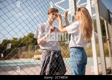 Ragazzo pensieroso che guarda sognatamente in macchina fotografica attraverso recinzione in rete mentre si trova di fronte a bella ragazza con capelli biondi. Giovane coppia che passa il tempo sopra Foto Stock