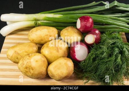 Patate giovani, ravanello, cipolle verdi, aneto su tavola di legno, fondo nero Foto Stock