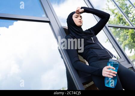 vista ad angolo basso della donna musulmana stanca in hijab con bottiglia sportiva vicino all'edificio moderno Foto Stock