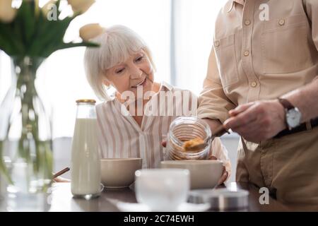 Fuoco selettivo di donna anziana sorridente seduta vicino l'uomo che versa i cereali in ciotola durante la colazione Foto Stock