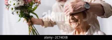 Scatto panoramico di uomo anziano che copre gli occhi alla moglie sorridente e che tiene il bouquet floreale a casa Foto Stock