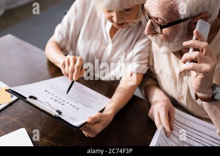 Vista ad alto angolo di un uomo anziano che parla su uno smartphone vicino alla moglie indicando un documento con la scritta di raccolta del debito Foto Stock