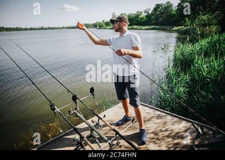 Pesca di pescatori giovani sul lago o sul fiume. Guy ha tre canne di pesca e cercando di trovare pesce in acqua. Uomo che lavora sulla regolazione della quarta asta. Pesca di alò Foto Stock