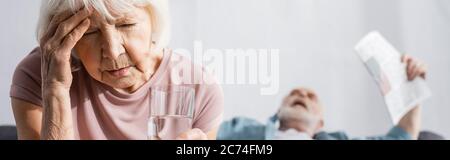 Foto panoramica di una donna anziana che tiene un bicchiere d'acqua vicino al marito con un giornale che soffre di calore a casa Foto Stock
