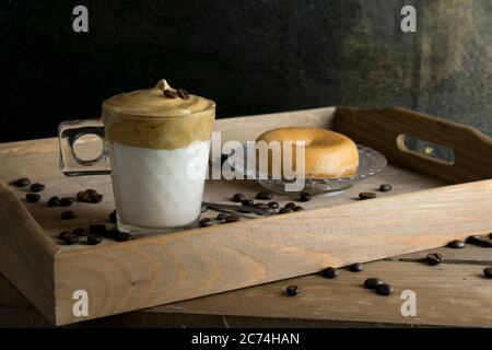 Montare il caffè istantaneo su un bicchiere di latte con una ciambella su un vassoio di legno con i chicchi di caffè spruzzati. Concetto di caffè Dalgona. Foto Stock