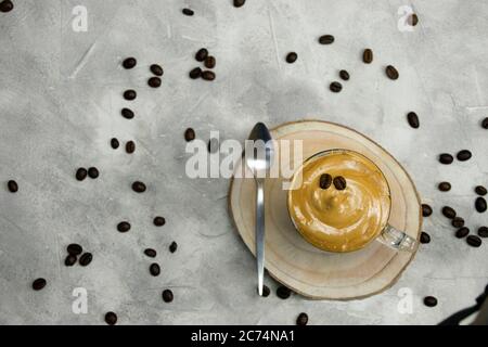 Vista dall'alto del caffè istantaneo montato con una tazza di cristallo con un cucchiaio su una montagne russe di legno su sfondo grigio con chicchi di caffè spruzzati e sfocati bac Foto Stock