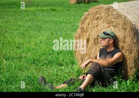 Seduto uomo al prato verde, basato su una balla di fieno, estate soleggiato giorno Foto Stock