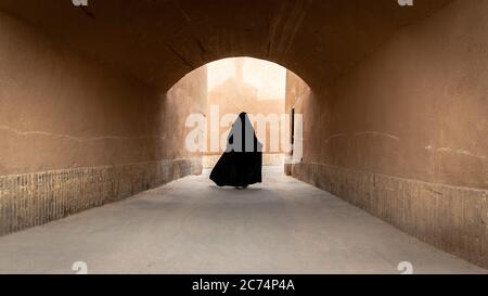 Donna musulmana non identificata, vestita in nero hijab cammina sulla strada stretta della città vecchia Yazd in Iran. Foto Stock