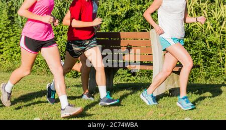 Un gruppo di ragazze delle scuole superiori si allenano insieme per correre in fondo sull'erba in un parco, passando una panchina del parco. Foto Stock