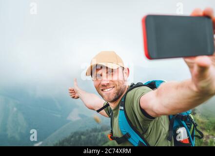 Giovane zaino in spalla uomo scattare selfie foto utilizzando smartphone e mostrare Thumbs Up durante la passeggiata dalla nebbia nuvolosa catena montuosa tempo. Attivo Foto Stock