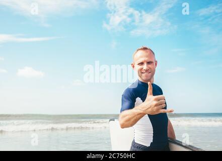 Ritratto di Surfer del giovane uomo che mostra il gesto famoso del segno di Shaka del surfer in macchina fotografica quando viene con una tavola da surf lunga a. onde Foto Stock