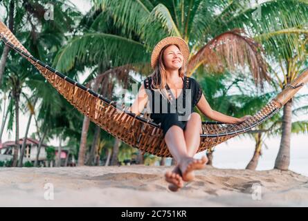 Giovane donna positiva seduta in amaca sulla spiaggia tropicale di palme